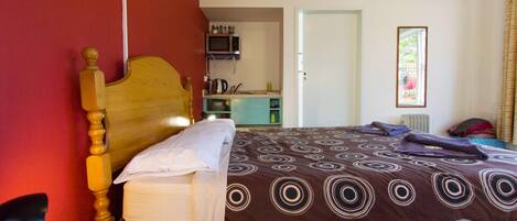 A room with a bed, a wooden headboard, a small table, a microwave, and a kitchenette in the background.