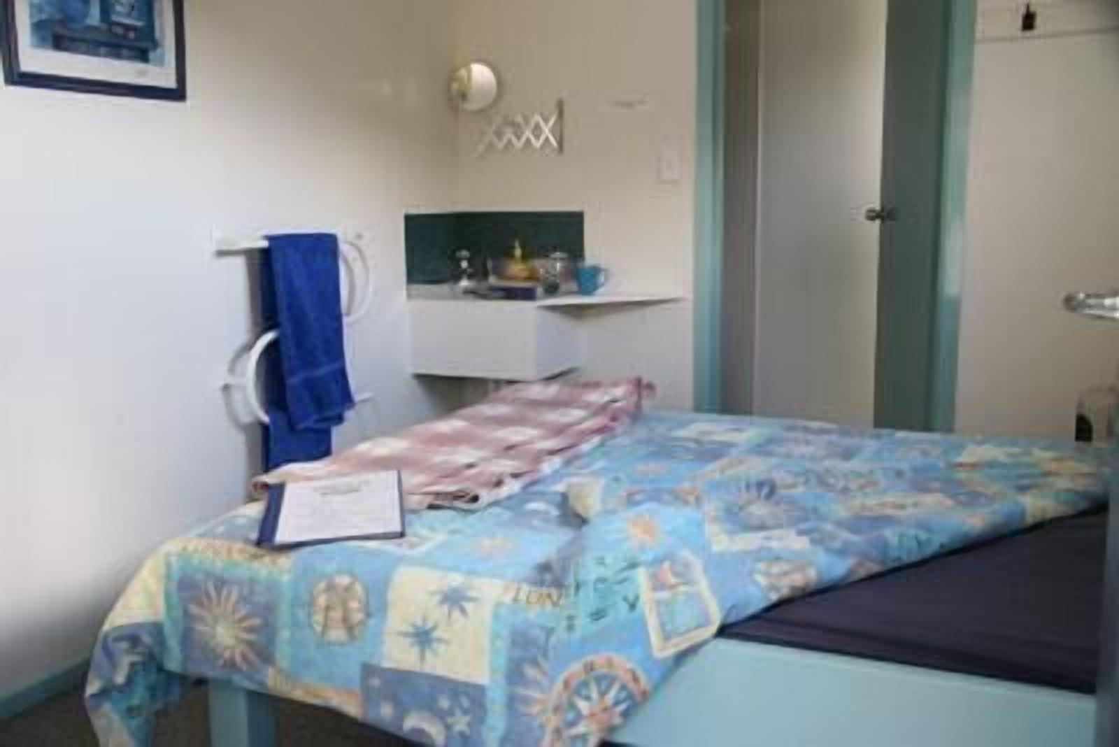 A small bedroom with a bed, a desk, and a chair. There is a towel hanging on the chair and a small shelf with items on it.