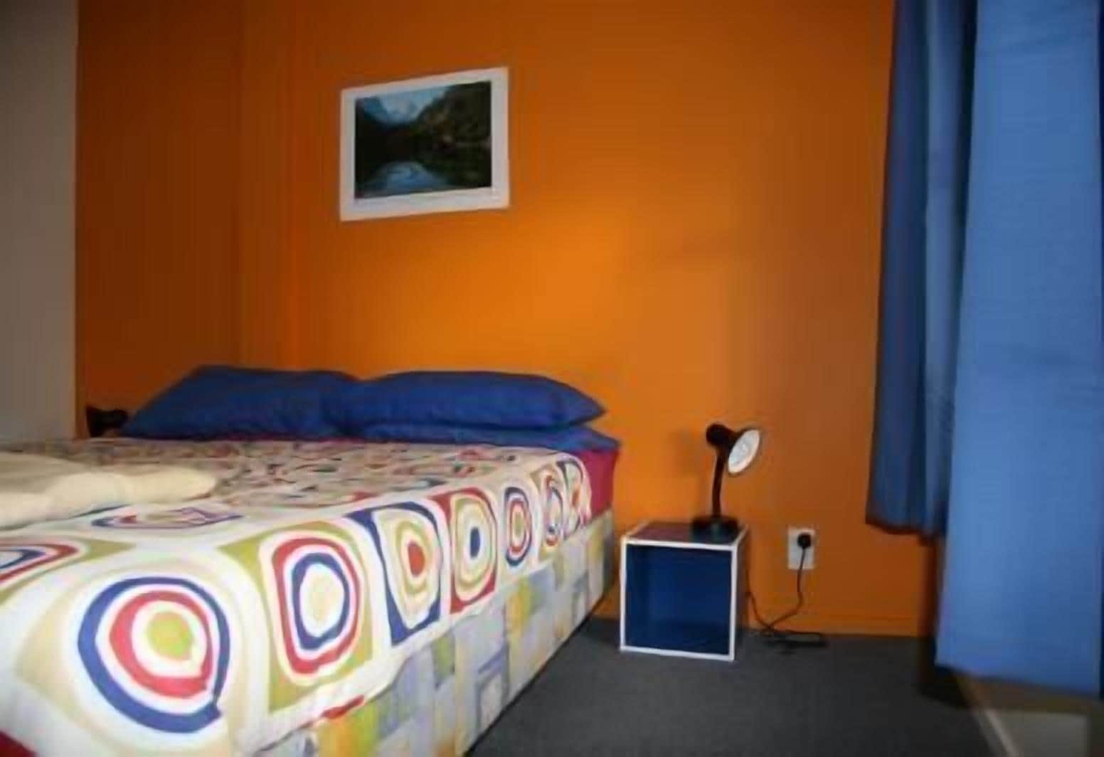 Ett rum med en orangefärgad vägg, en säng med ett färgglatt täcke, ett litet skrivbord med en lampa och en tavla på väggen.