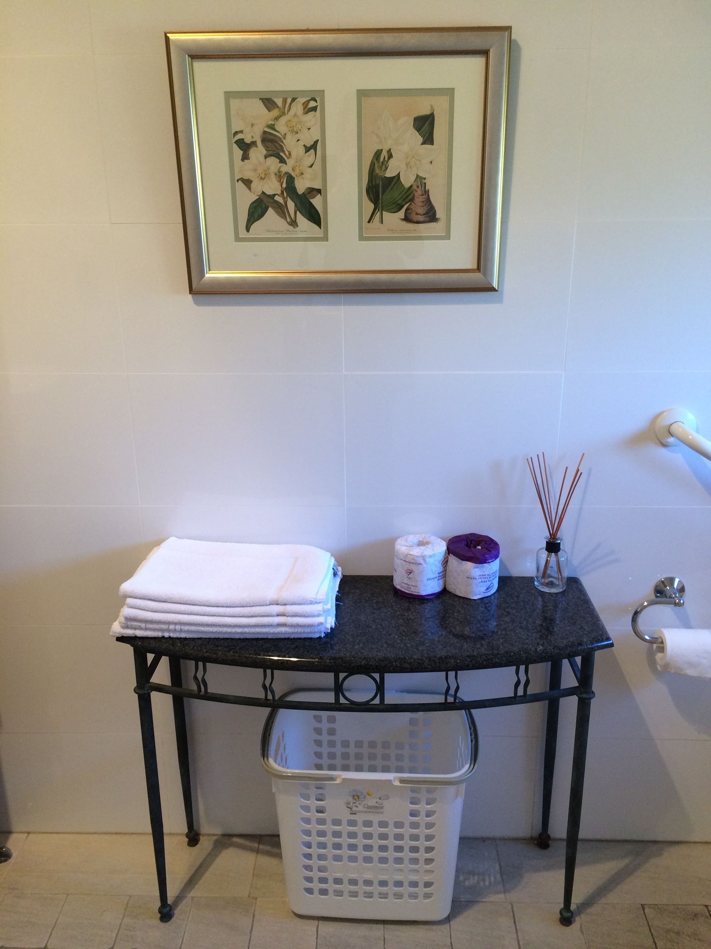 Une salle de bain avec un mur carrelé, une illustration botanique encadrée, une pile de serviettes blanches, une petite table avec un flacon de désodorisant, une bouteille en verre et un panier à linge.