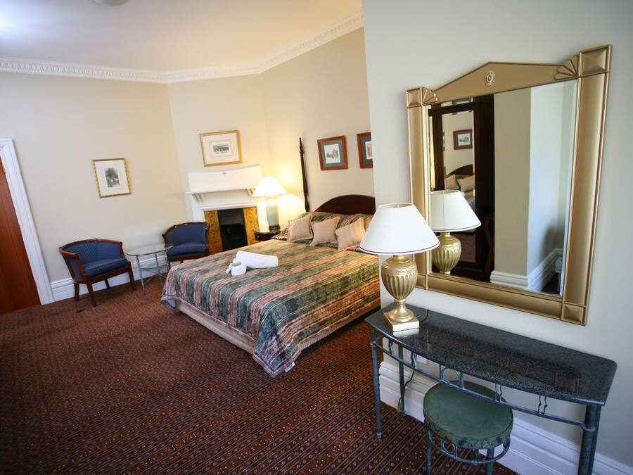 A hotel room with a bed, two chairs, a desk with a lamp, a mirror, and framed pictures on the wall.