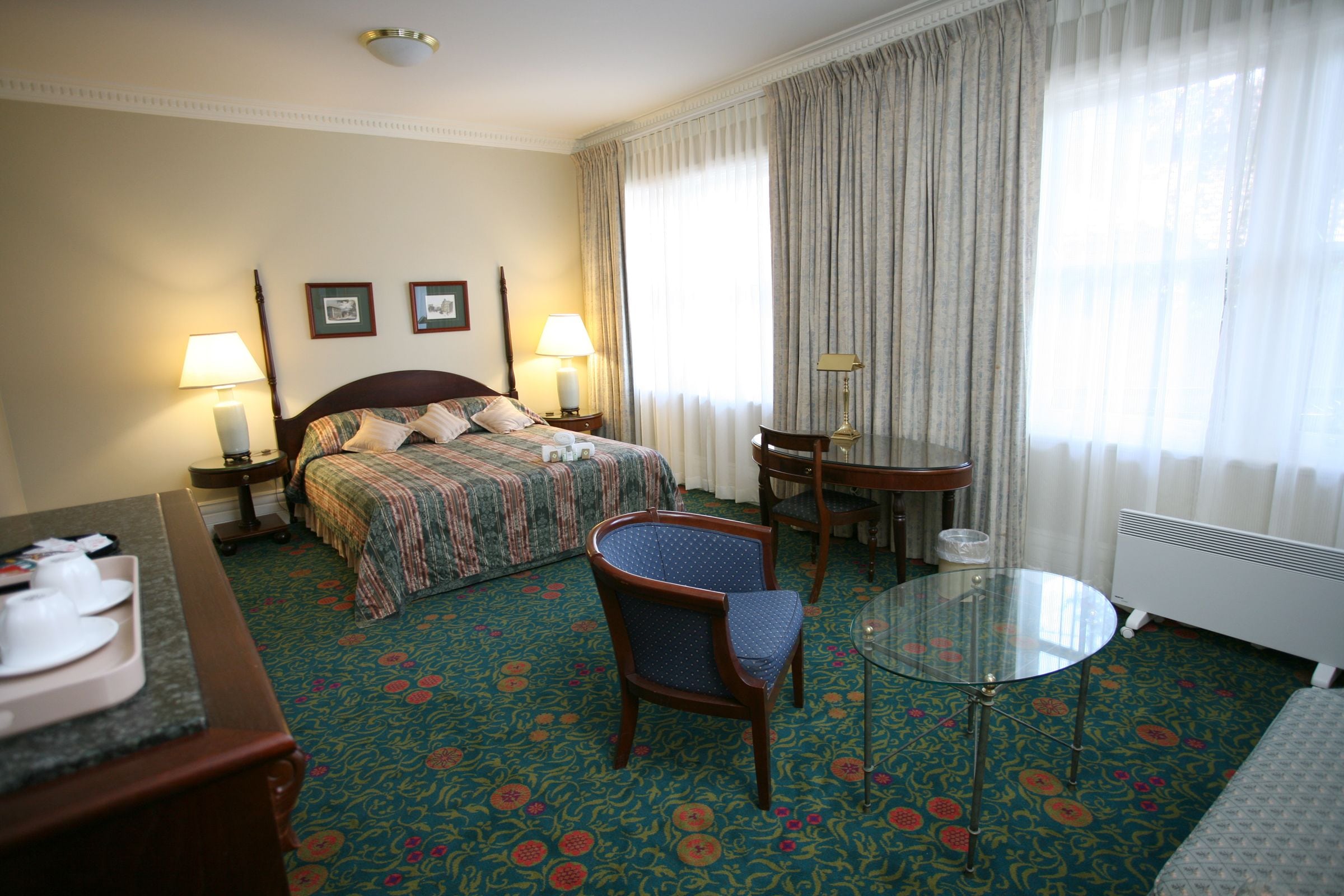 A hotel room with a bed, two bedside lamps, a chair, a small table, and a radiator.