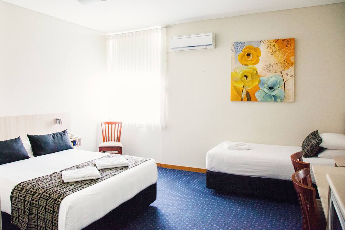 Quarto de hotel com duas camas, uma cadeira, uma escrivaninha e um quadro na parede.