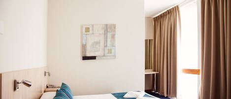 Een hotelkamer met een bed, nachtkastje, lamp, schilderij aan de muur en een raam met gordijnen.