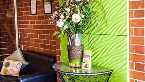 A room with a brick wall, a green textured partition, a black leather sofa, a small round table with a vase of flowers, and an air conditioning unit mounted on the wall.