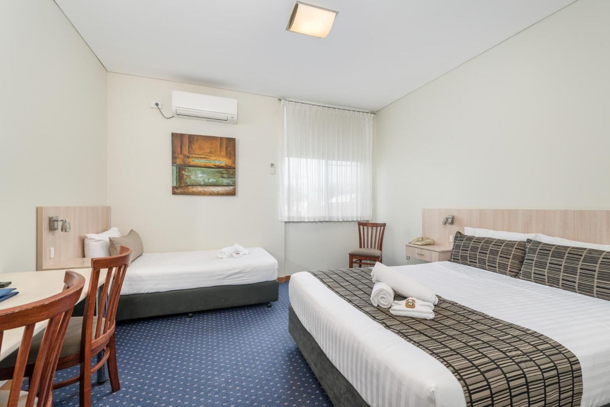 A hotel room with two single beds, a desk, a chair, a small table, a telephone, and a painting on the wall.