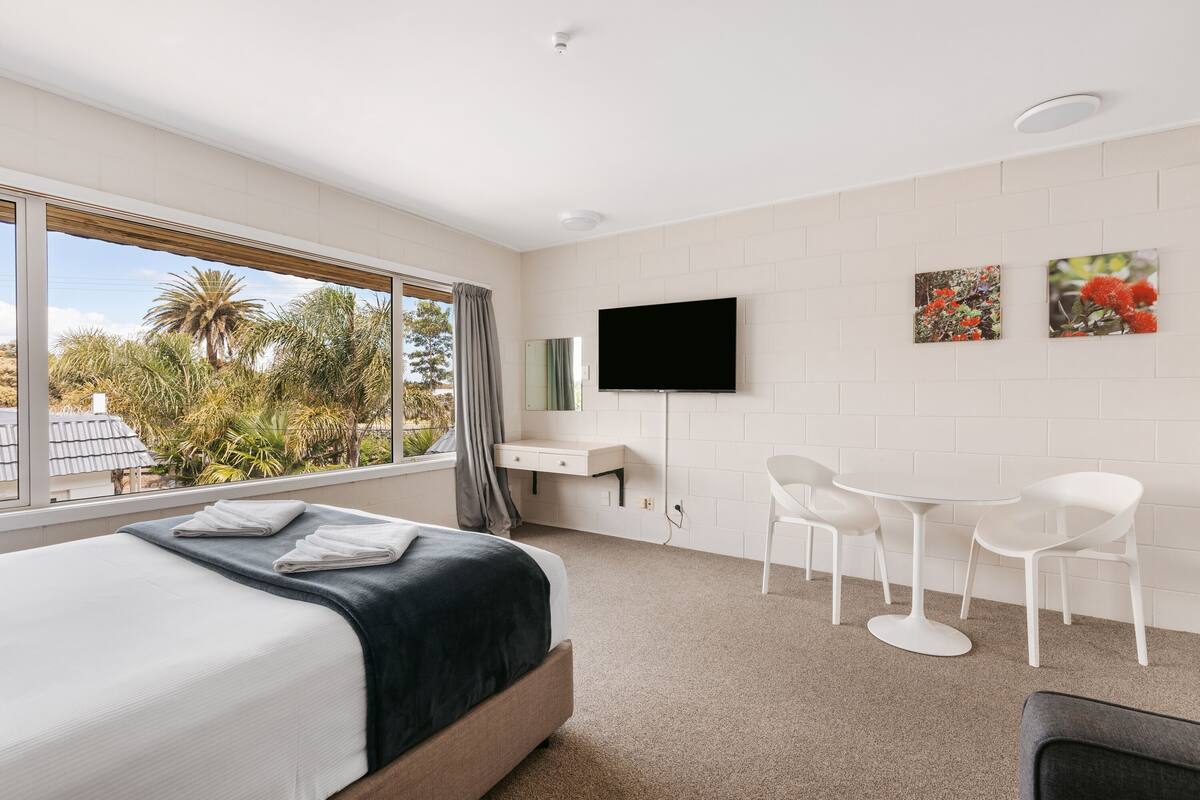 A modern hotel room with a large bed, a desk, two chairs, a TV, and a window with a view of palm trees.