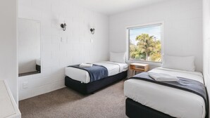 A hotel room with two beds, a small table, a window with a view of trees, and white brick walls. - Avalon Motel Thames – Wenzel Motels (Thames)