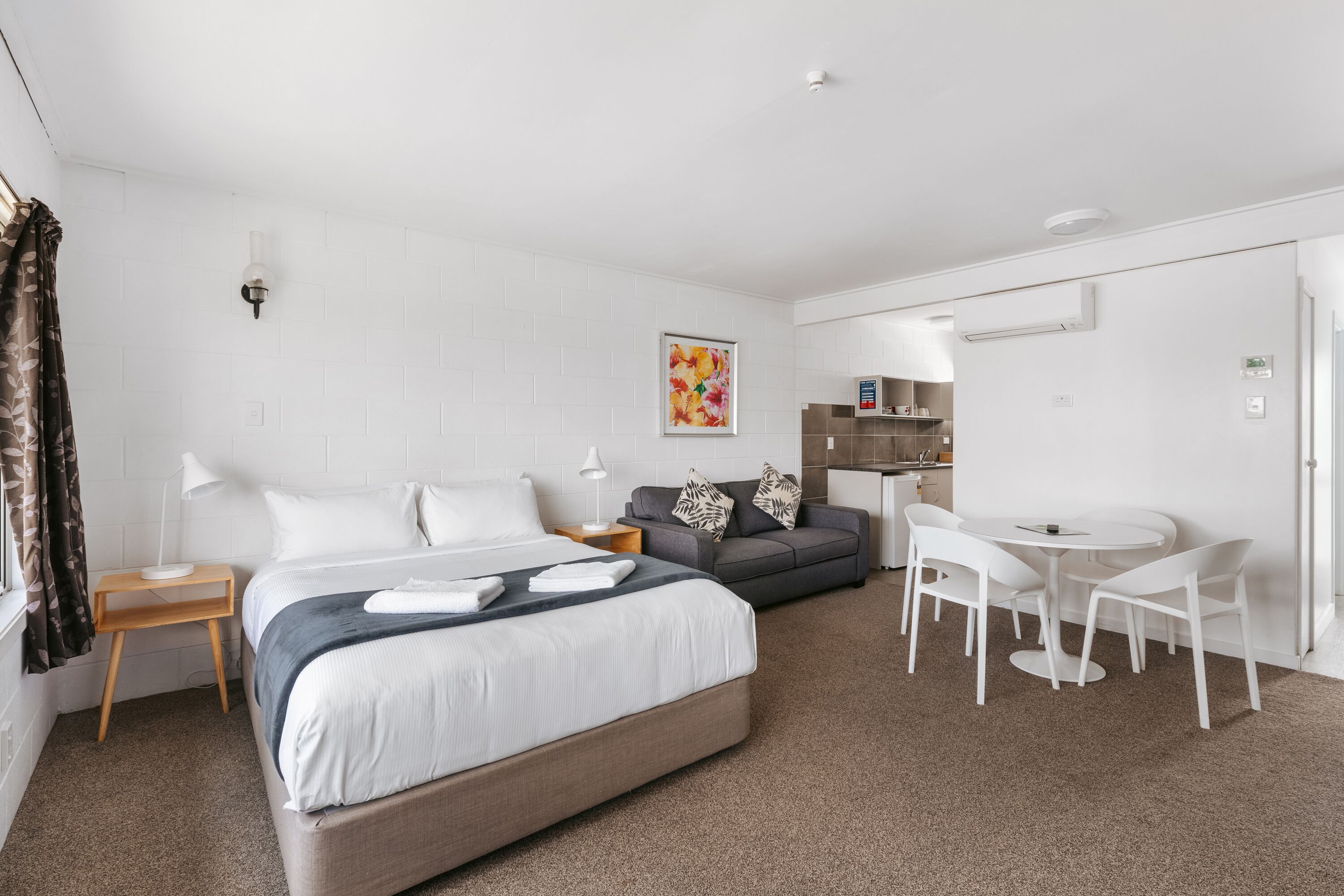 A modern hotel room with a large bed, a sofa, a dining table with chairs, a small side table, and a wall-mounted air conditioning unit.
