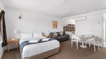 A modern hotel room with a large bed, a sofa, a dining table with chairs, a small side table, and a wall-mounted air conditioning unit.