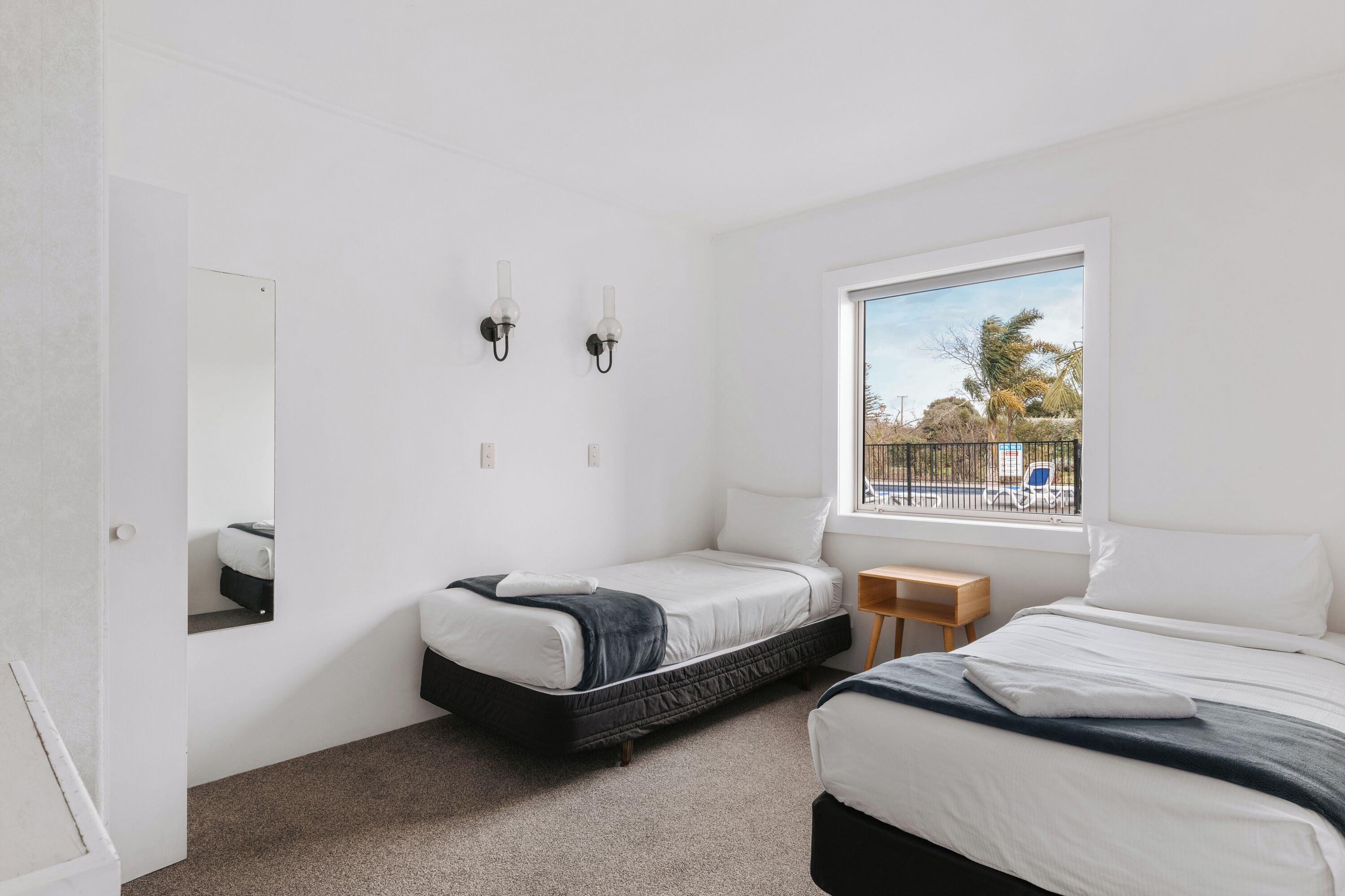 A bedroom with two beds, a window with a view, a mirror, and a small wooden table.