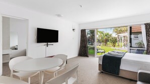 A hotel room with a bed, a TV, a dining table with chairs, and a sliding glass door leading to a garden. - Avalon Motel Thames – Wenzel Motels (Thames)