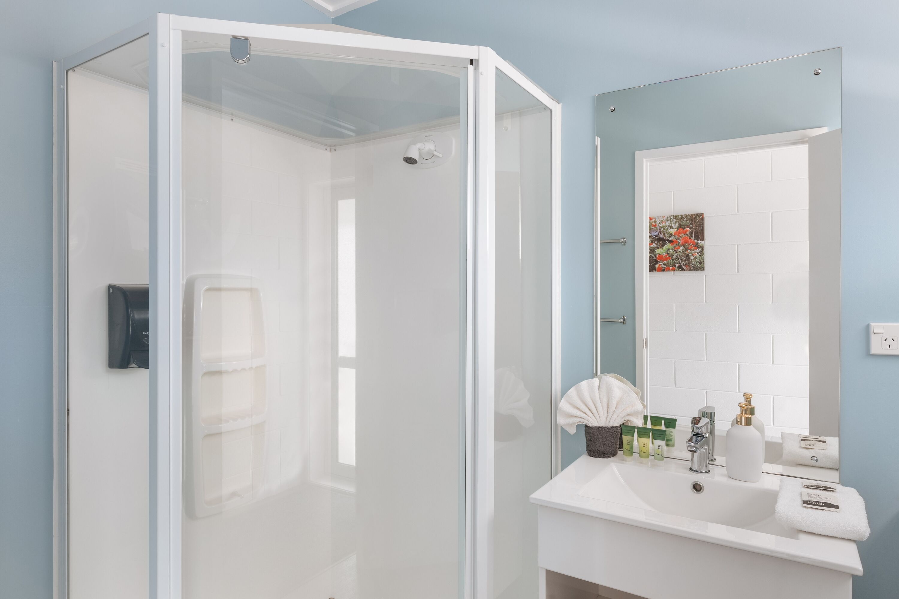 A modern bathroom with a glass shower enclosure, a white sink with toiletries, and a mirror with a floral artwork.
