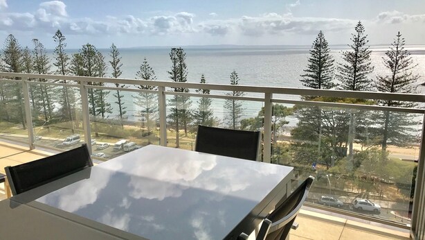 A balcony with a table and chairs, overlooking a beach and trees. - Proximity Waterfront Apartments (Redcliffe)