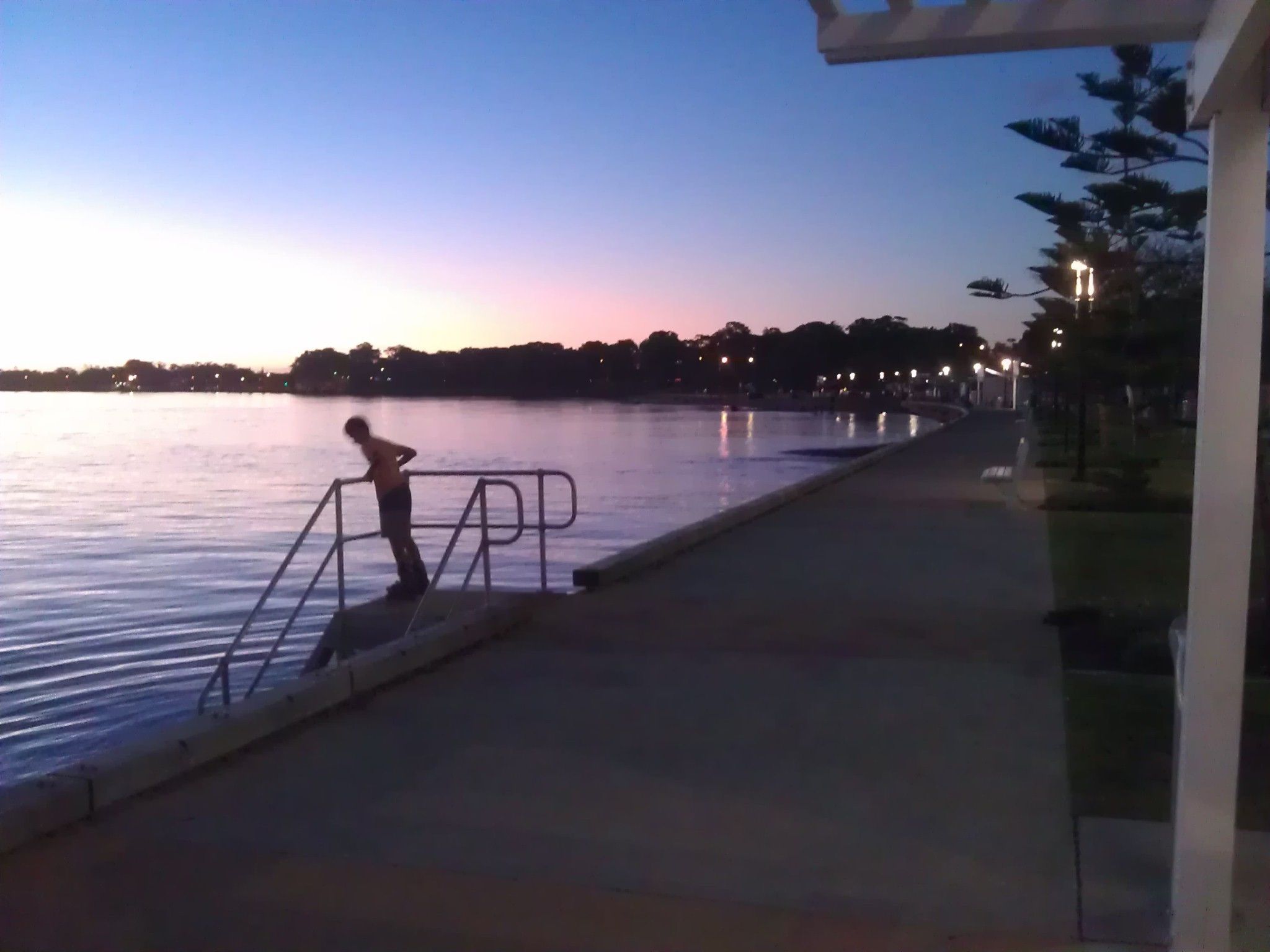 A waterfront walkway with a metal railing, a person standing by the railing, and a calm body of water reflecting lights from the background.