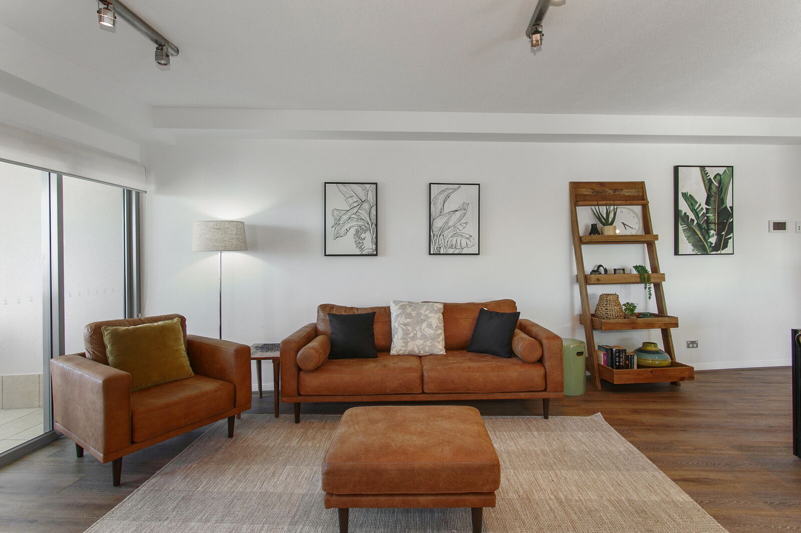 A modern living room with a brown leather sofa, a matching ottoman, and a wooden side table. There are framed botanical prints on the wall and a wooden plant stand with books and decorative items.