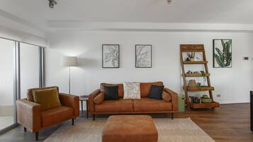 A modern living room with a brown leather sofa, a matching ottoman, and a wooden side table. There are framed botanical prints on the wall and a wooden plant stand with books and decorative items.