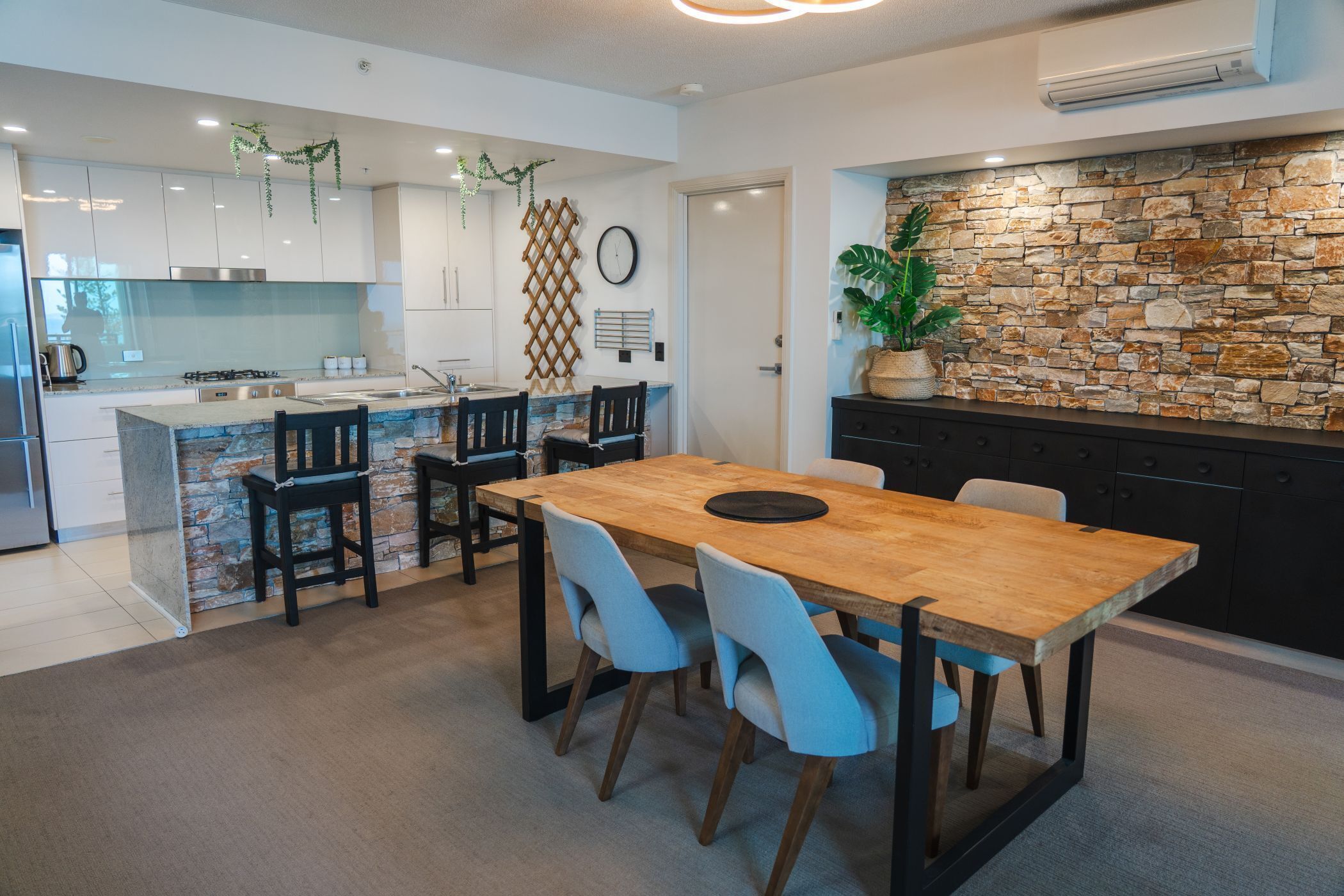 Een moderne keuken met een achterwand van steen, een houten eettafel met stoelen en een inbouwkookeiland met barkrukken.