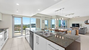 A modern kitchen with a large island, a dining area with a table and chairs, a grey sofa, and a TV. - Proximity Waterfront Apartments (Redcliffe)