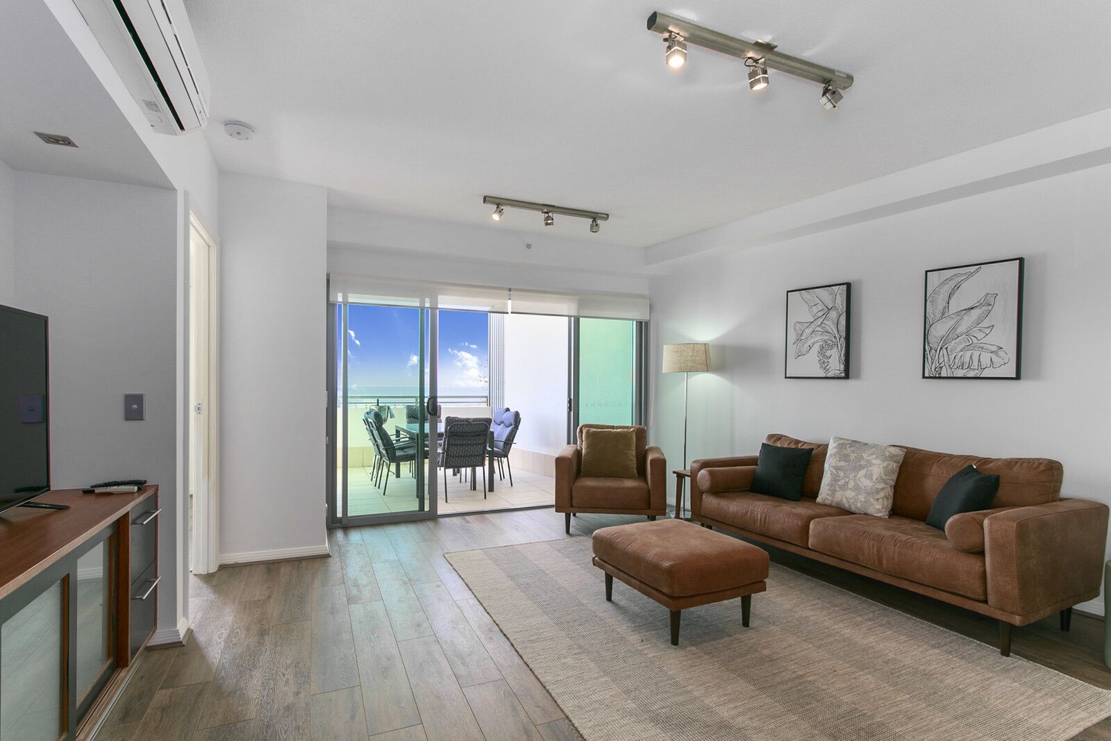 Ein modernes Wohnzimmer mit einer braunen Ledersofa, einem Glastisch mit Stühlen, einem Flachbildfernseher und einer großen Schiebeglastür, die auf einen Balkon mit Blick auf den Himmel und die Wolken führt.