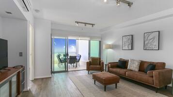A modern living room with a brown leather sofa, a glass dining table with chairs, a flat-screen TV, and a large sliding glass door leading to a balcony with a view of the sky and clouds.