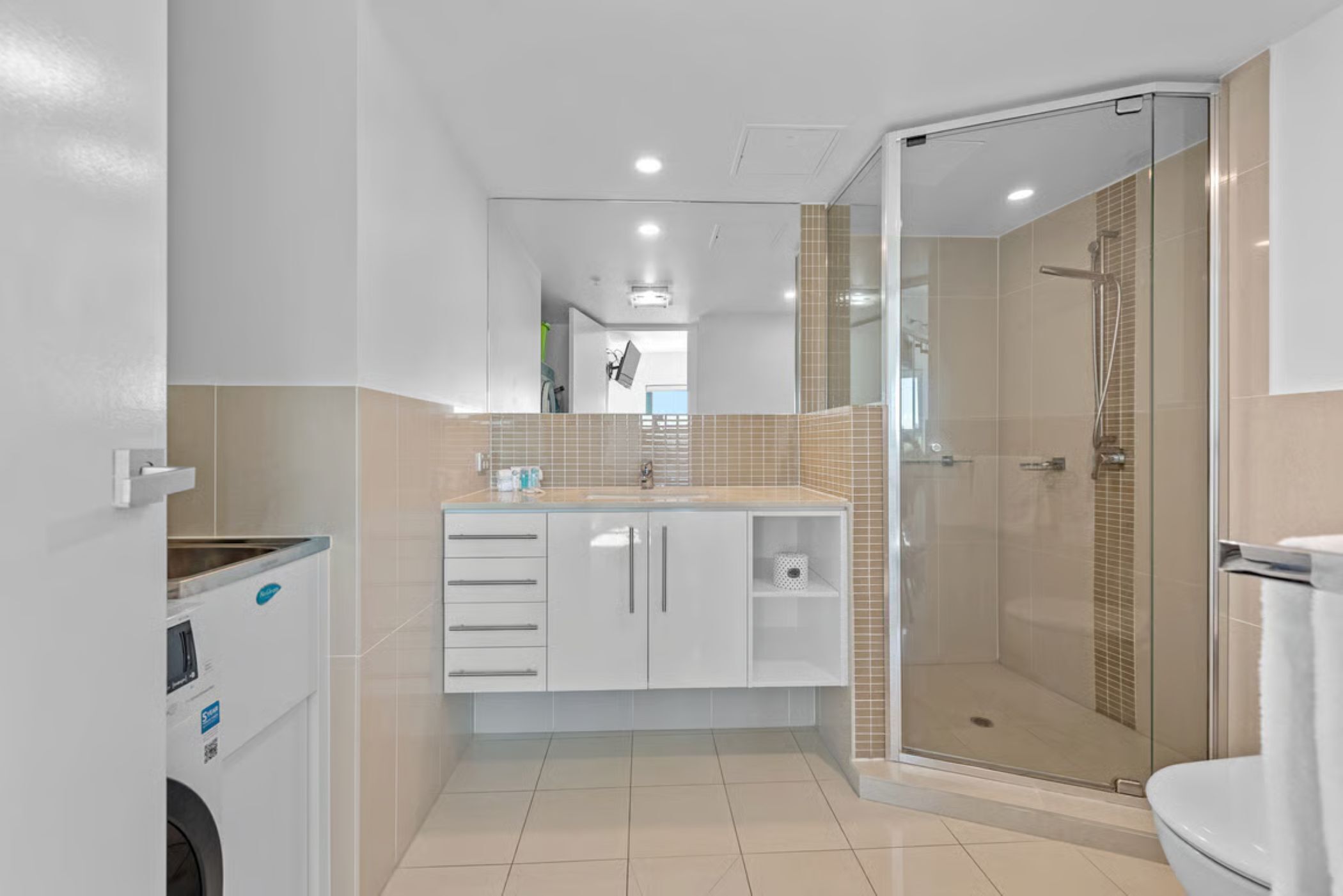 A modern bathroom with a glass shower enclosure, a white sink with a mirror above, and a built-in washing machine.