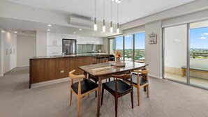 A modern kitchen with a central island, a dining area with a wooden table and chairs, and large windows offering a city view. - Proximity Waterfront Apartments (Redcliffe)