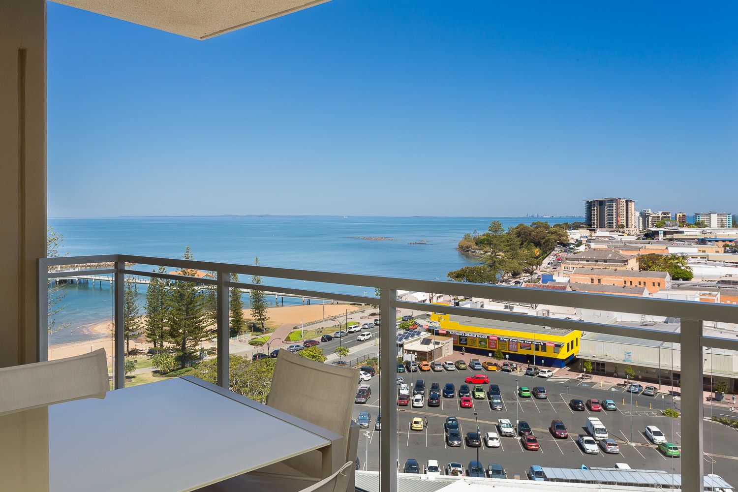 A balcony with a view of a parking lot, a yellow bus, and a beach with a pier.