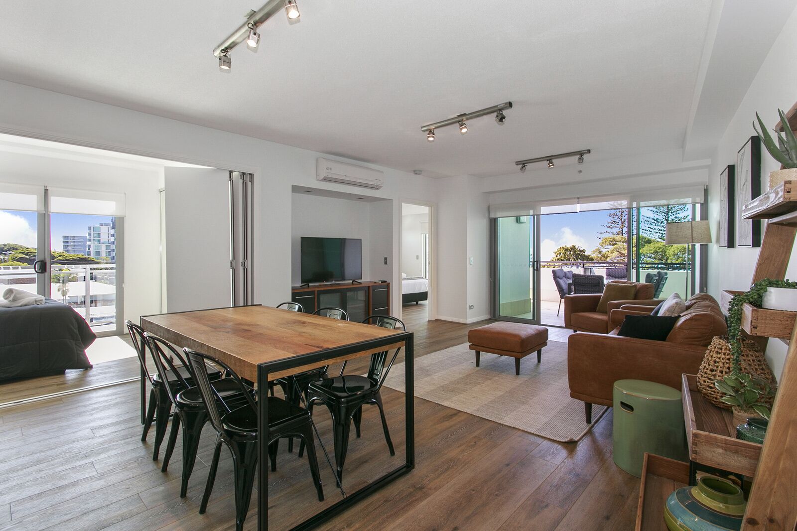 A spacious living area with a wooden dining table, black chairs, a sofa, and a green ottoman. There is a television and a balcony with a view of the city.