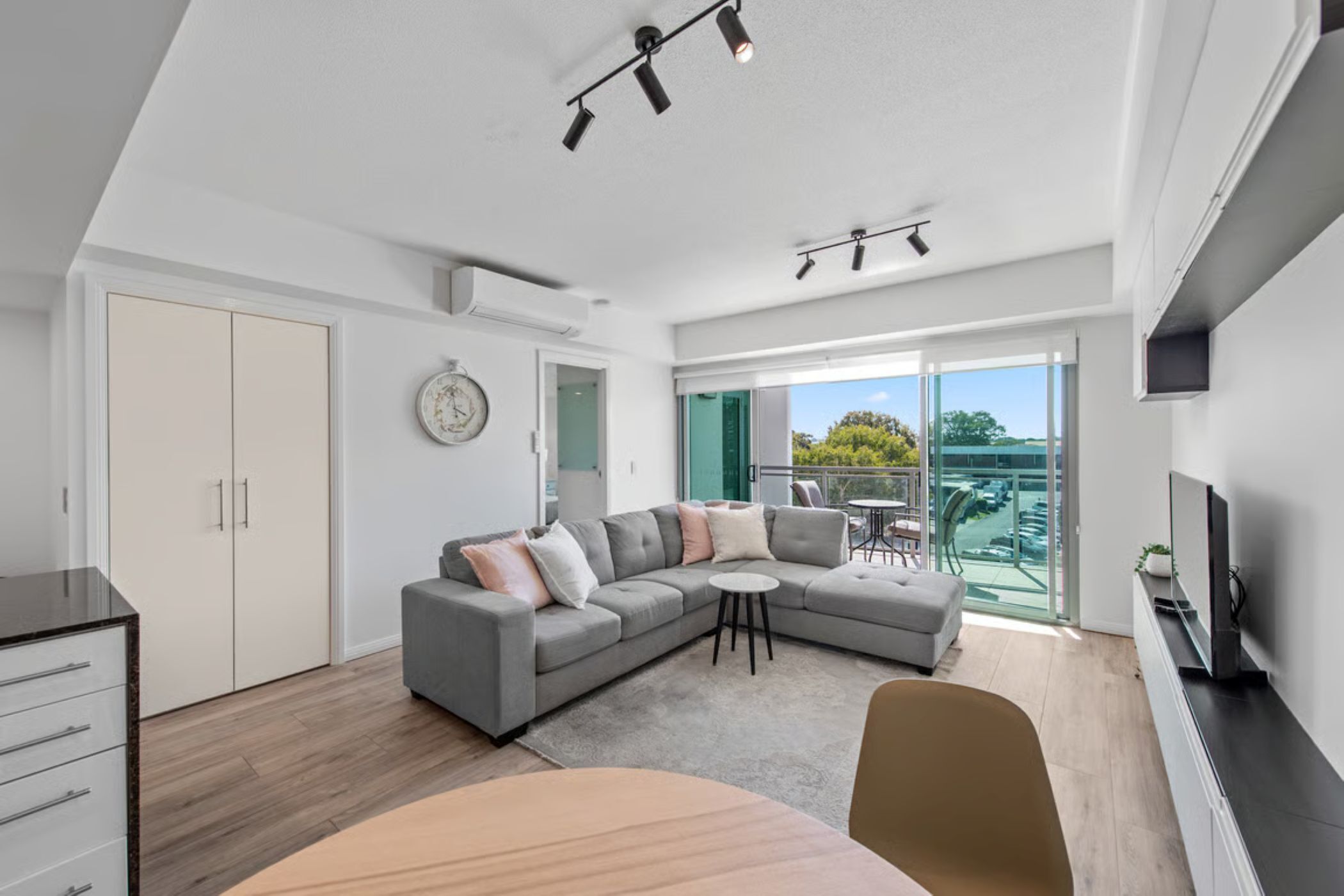A modern living room with a grey sectional sofa, a round coffee table, and a TV on a black stand. There is a large window with a view of greenery and a balcony.