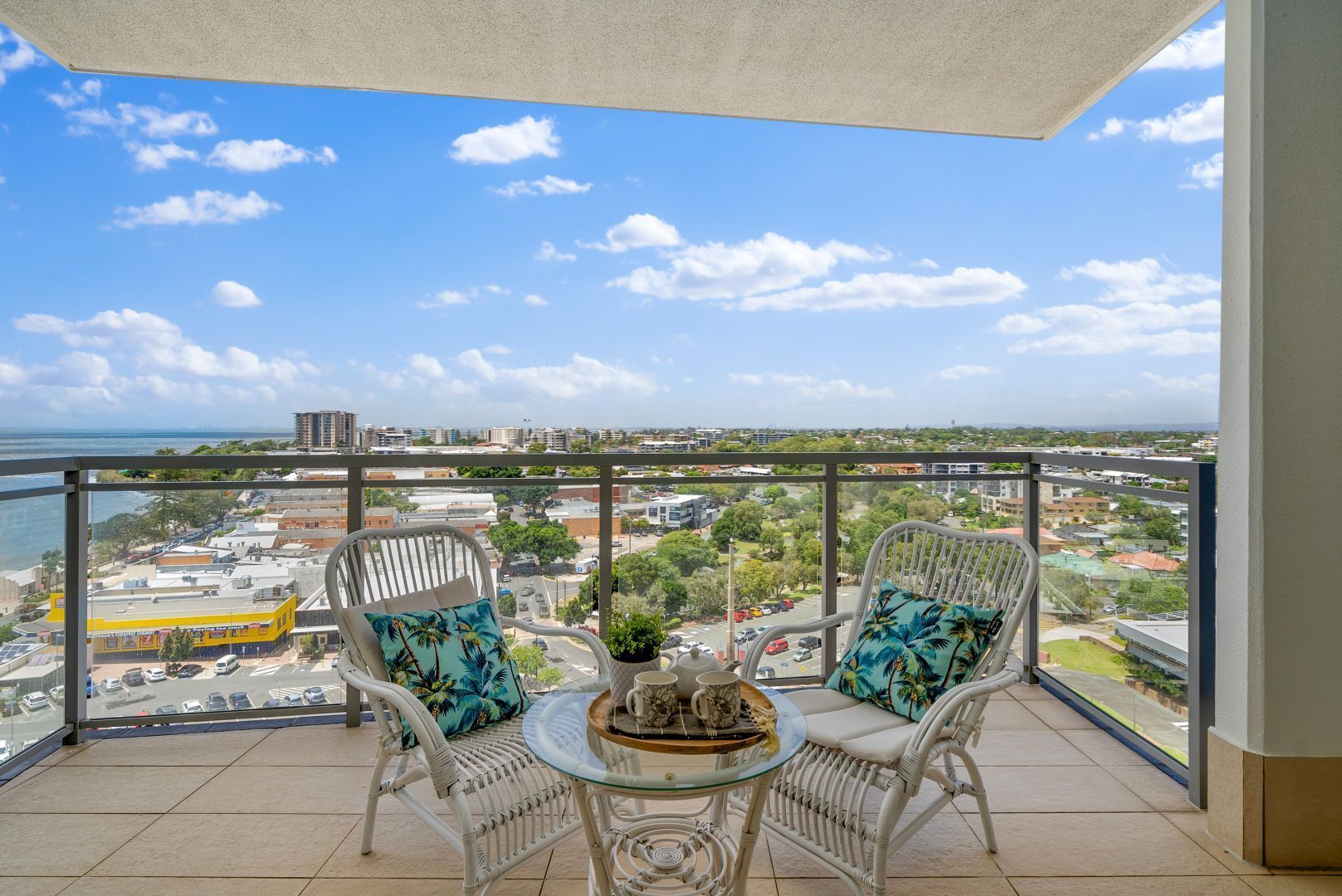 A balcony with two wicker chairs, a glass table, and a view of a cityscape and body of water.