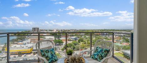 A balcony with two wicker chairs, a glass table, and a view of a cityscape and body of water.