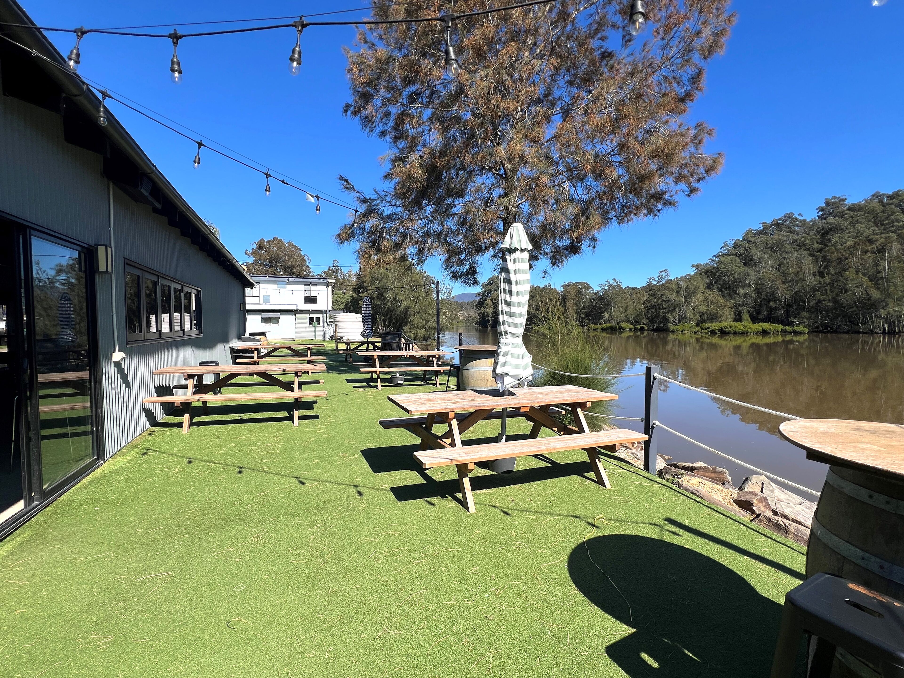 An outdoor patio with picnic tables and string lights, overlooking a calm lake and surrounded by trees.