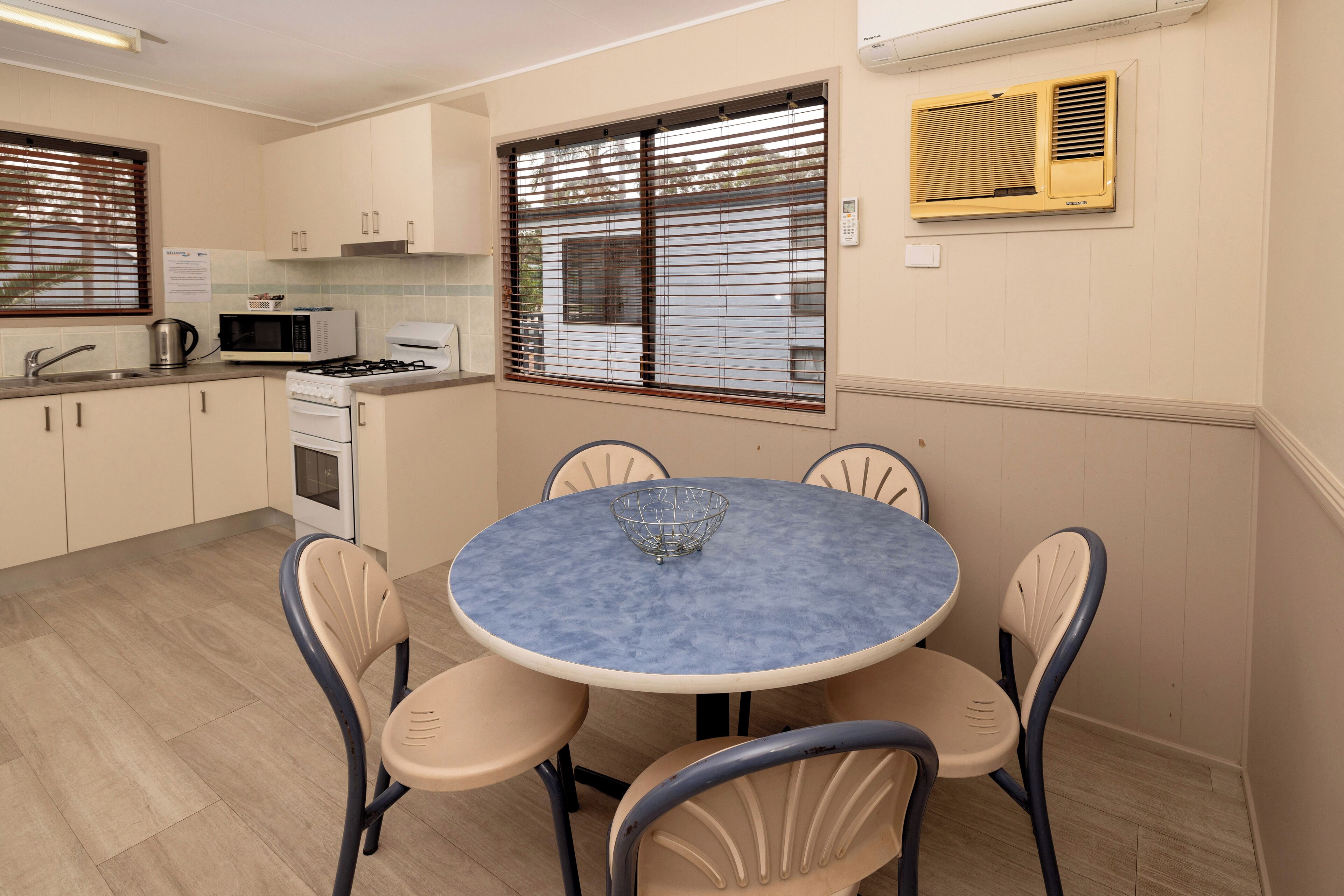 Cocina compacta con estufa a gas, microondas y pileta. Hay una mesa redonda con cuatro sillas y una ventana con persianas.
