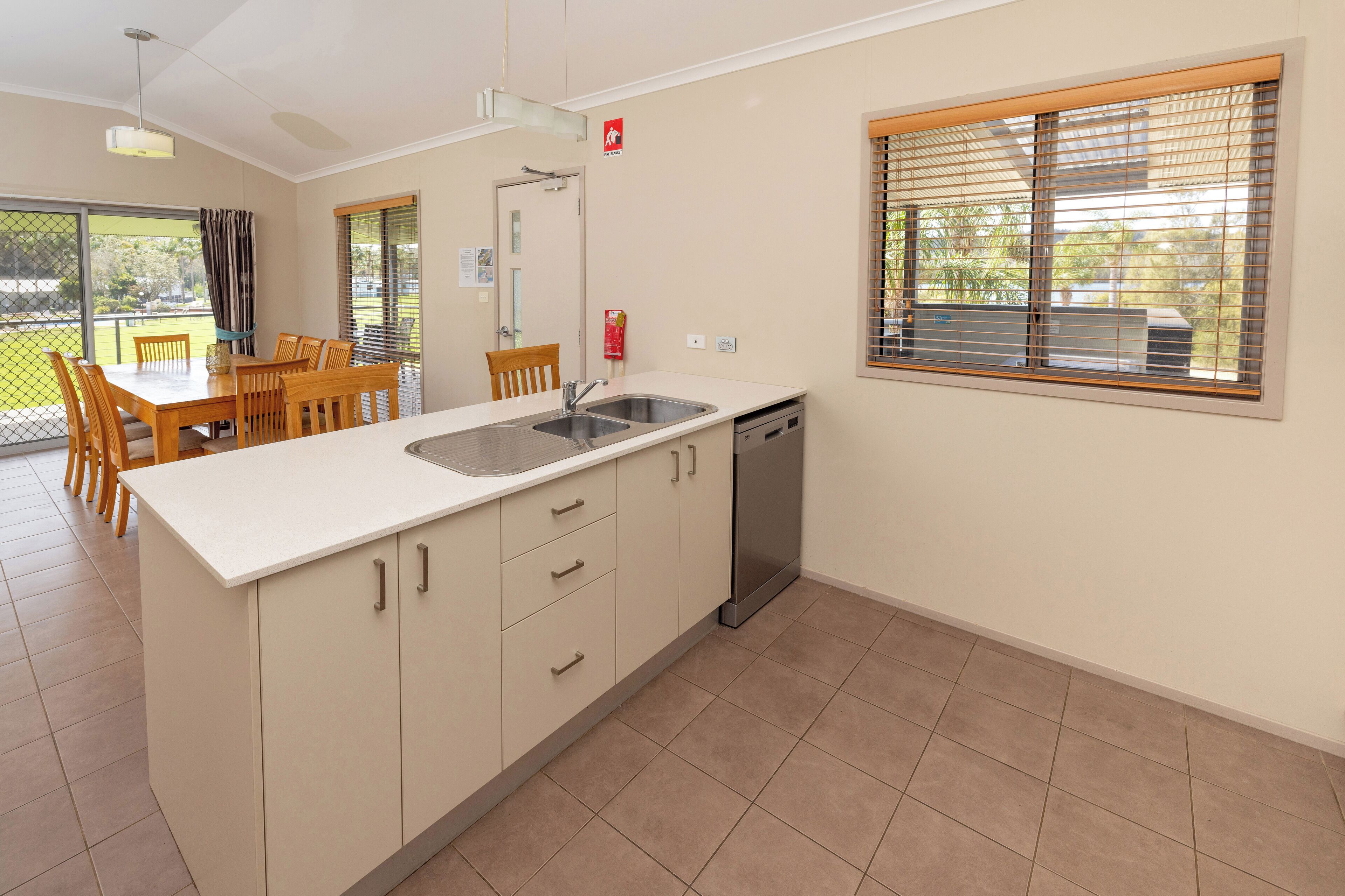 Una cocina moderna con encimera blanca, pileta de acero inoxidable y lavavajillas. Hay una zona de comedor con sillas de madera y una mesa.