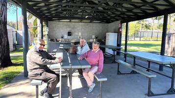 Un'area picnic coperta con panche e tavoli, un pavimento in cemento e un edificio con una cucina sullo sfondo.