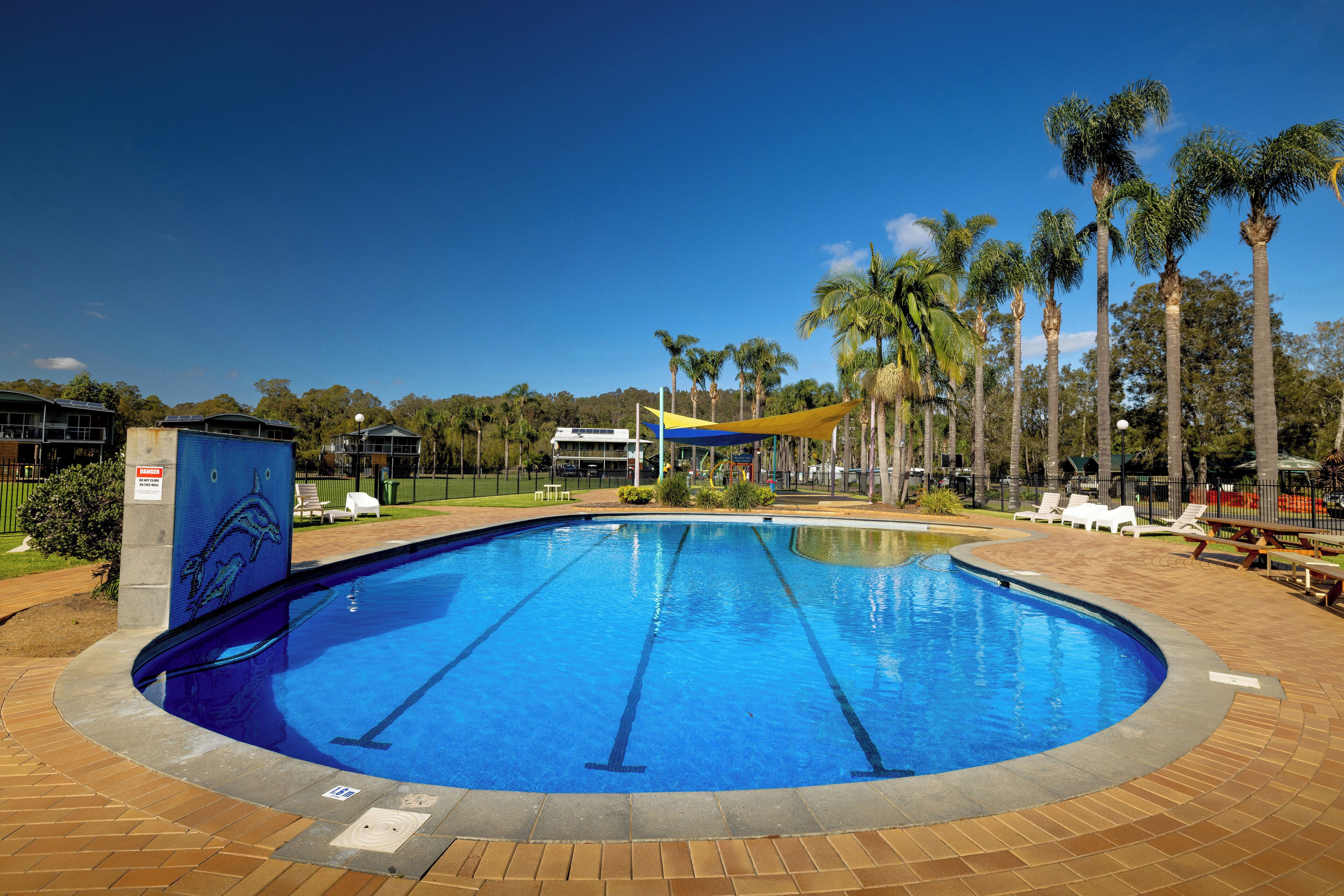 Piscina com água azul cristalina, rodeada por um deck de tijolos e palmeiras.