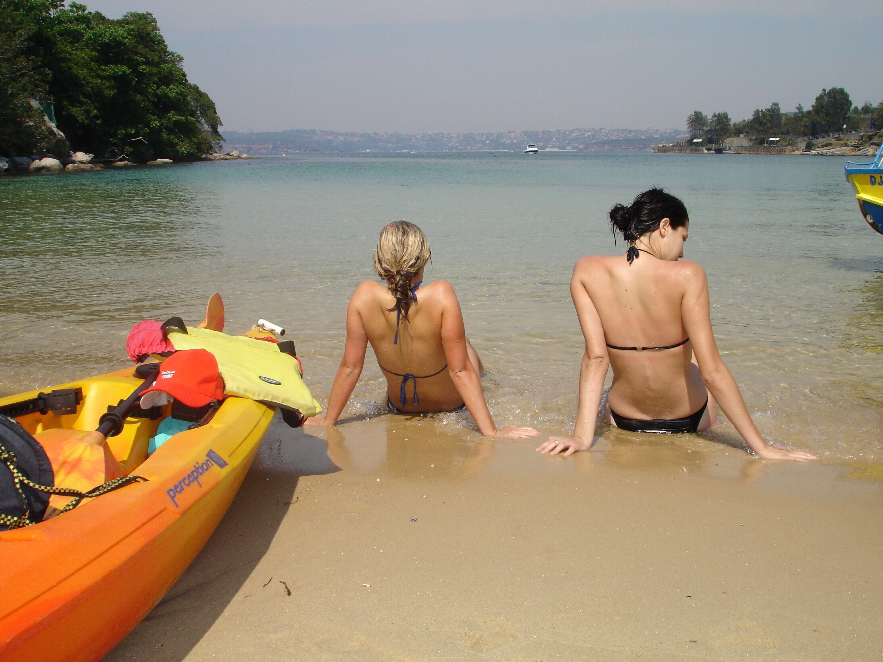 Dos mujeres en trajes de baño en una playa, con un bote amarillo y una costa rocosa al fondo.