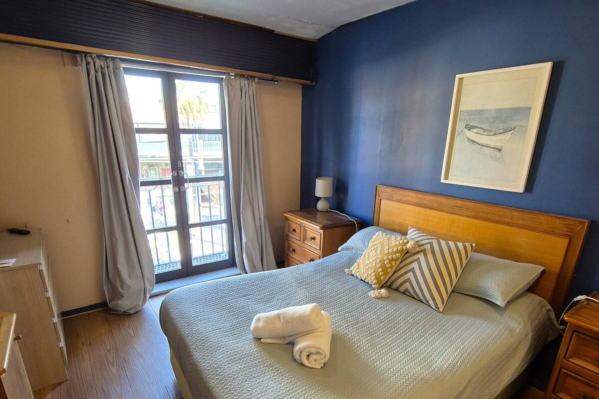 A neatly made bed with pillows and a rolled towel, wooden furniture, a window with curtains, and a painting on the wall.