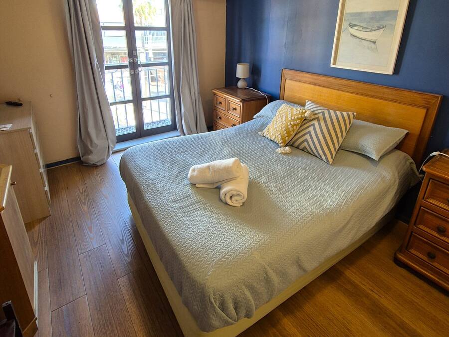 Una cama bien hecha con almohadas y una toalla enrollada, muebles de madera, una ventana con cortinas y un cuadro en la pared.