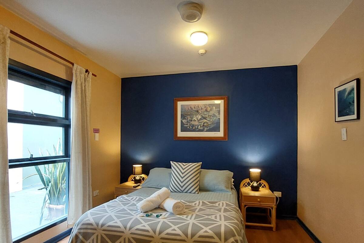 A bedroom with a bed, two bedside tables, a window, a painting on the wall, and a ceiling light.