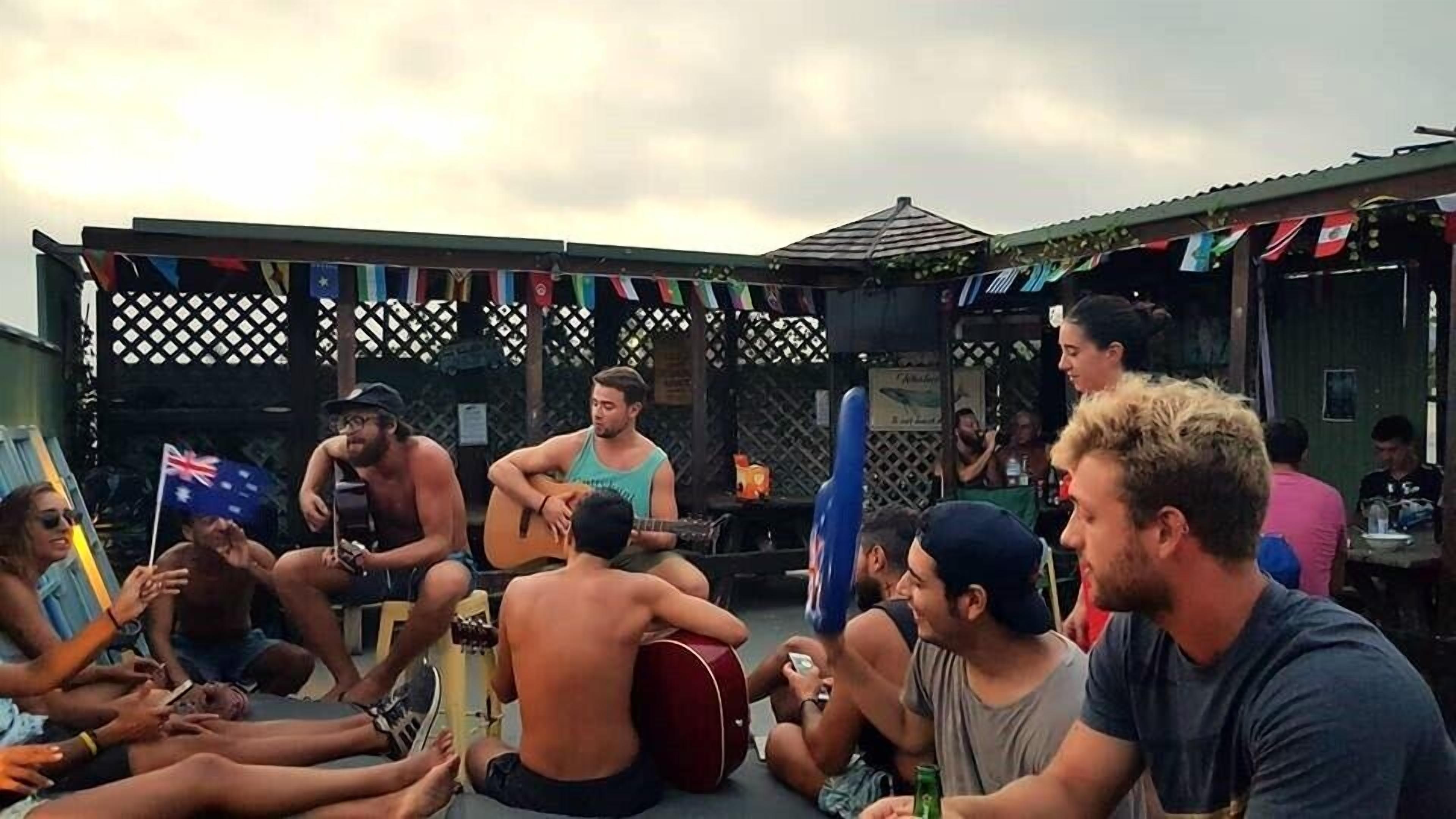 Un groupe de personnes est assis à l’extérieur, certaines jouent de la guitare, et on aperçoit une clôture et des drapeaux à l’arrière-plan.