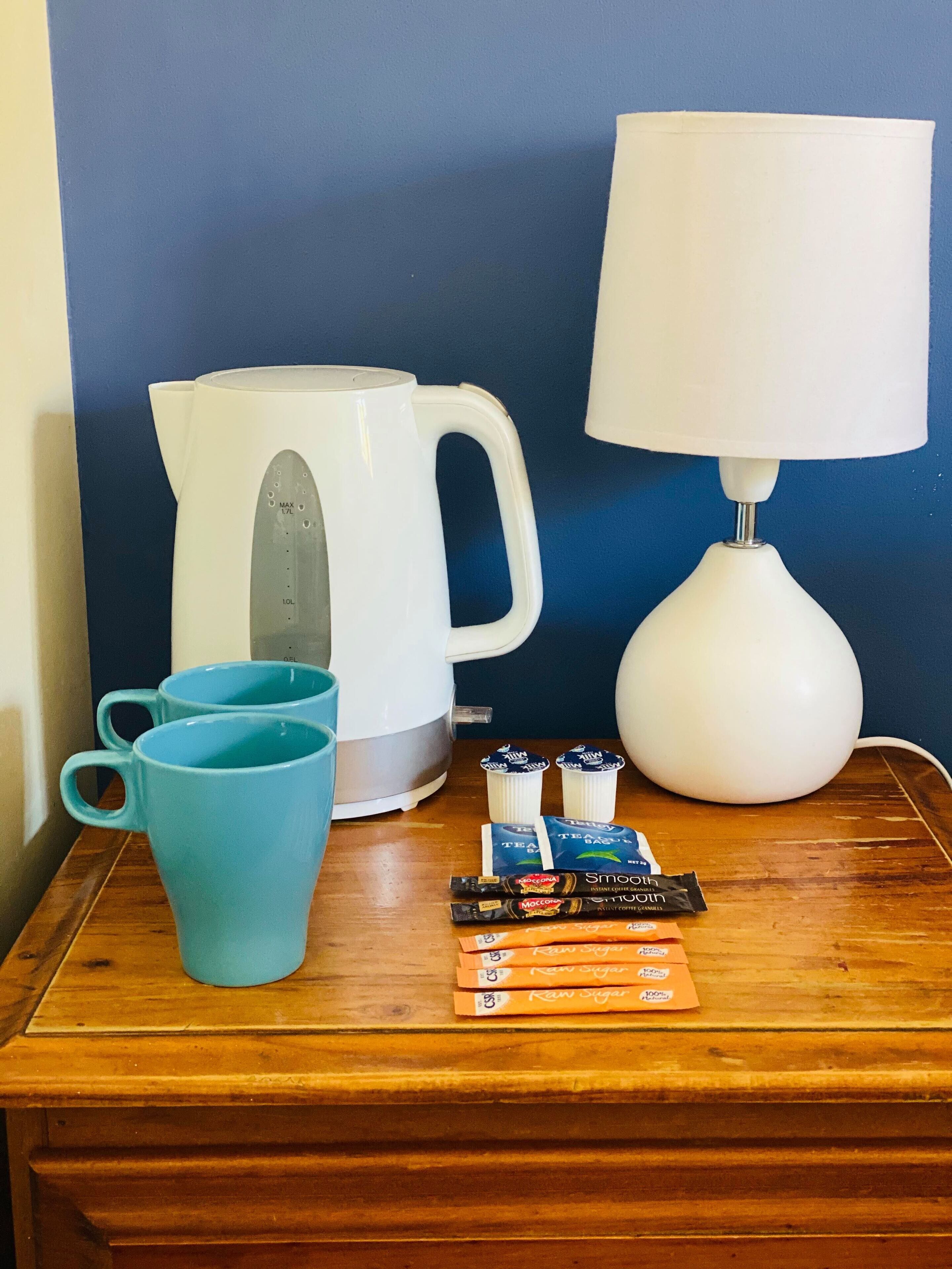 Una mesita de noche de madera con una tetera blanca, una taza azul, una lámpara blanca y algunos paquetes de té y café.