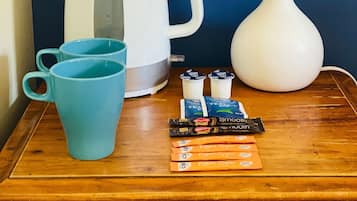 Una mesita de noche de madera con una tetera blanca, una taza azul, una lámpara blanca y algunos paquetes de té y café.