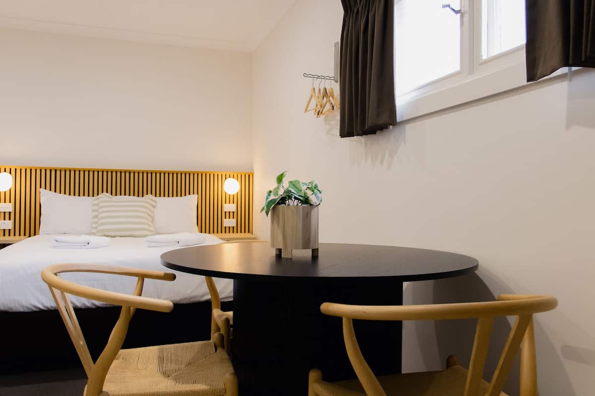 A hotel room with a bed, a round table, two wooden chairs, a potted plant, and a window with curtains.