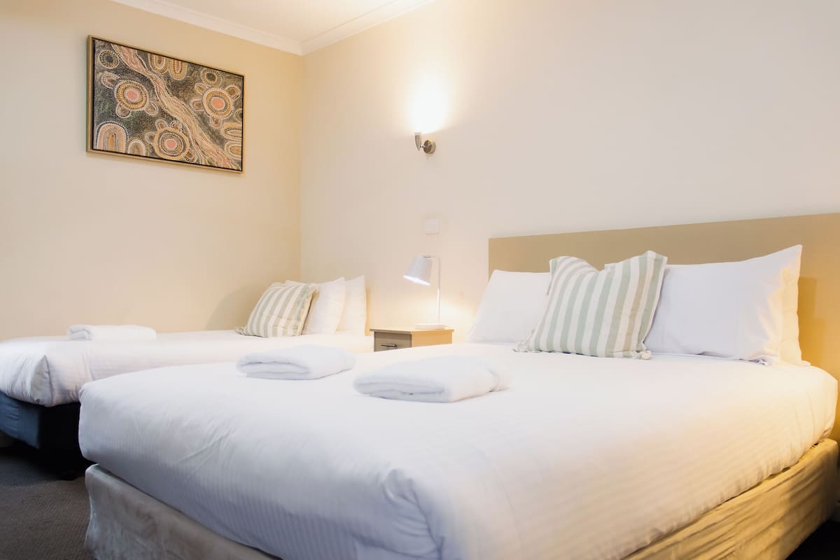 A hotel room with two beds, each with white linens and pillows, a bedside table with a lamp, and a wall-mounted artwork.