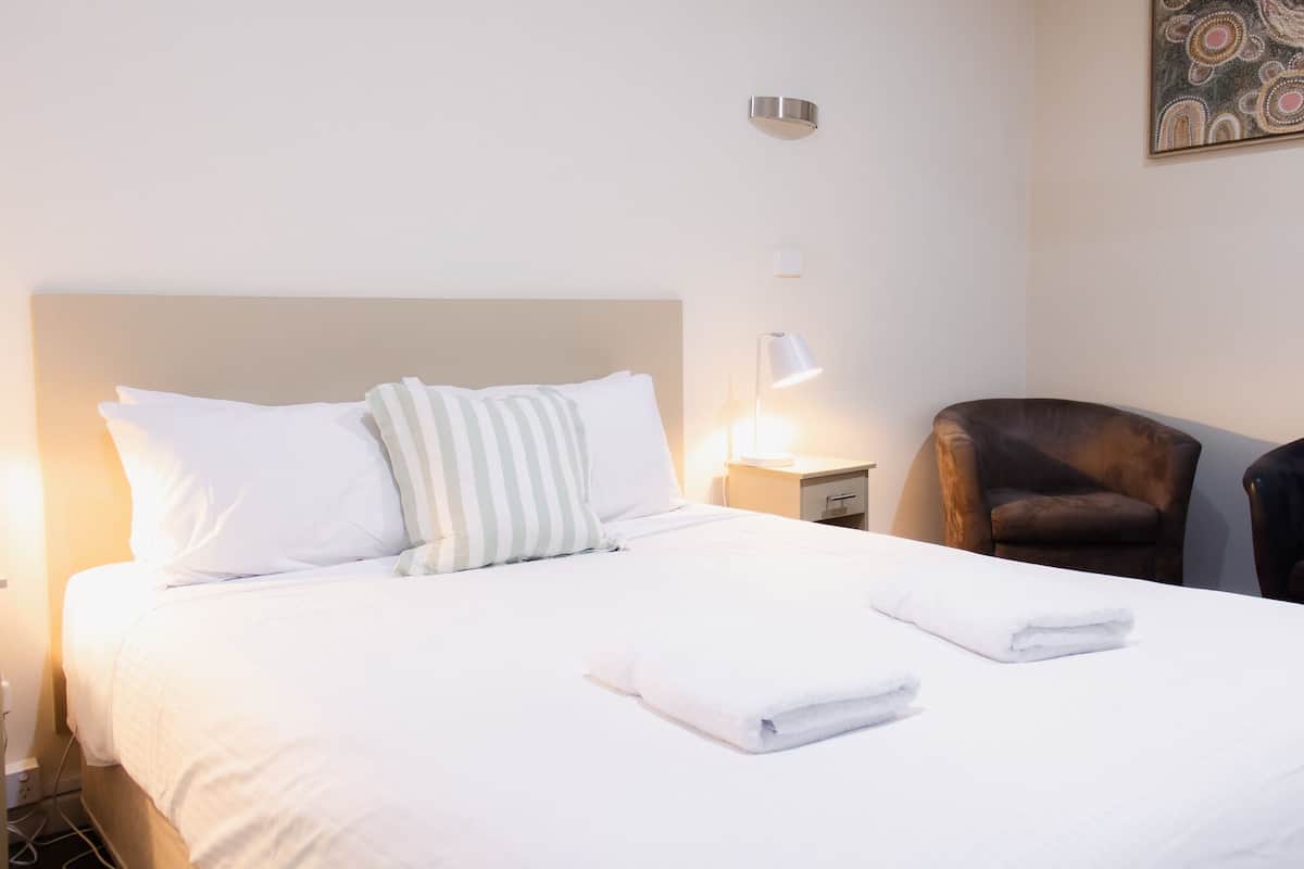 A neatly made bed with white linens, a striped pillow, and two folded towels. There is a brown leather chair and a small bedside table with a lamp.