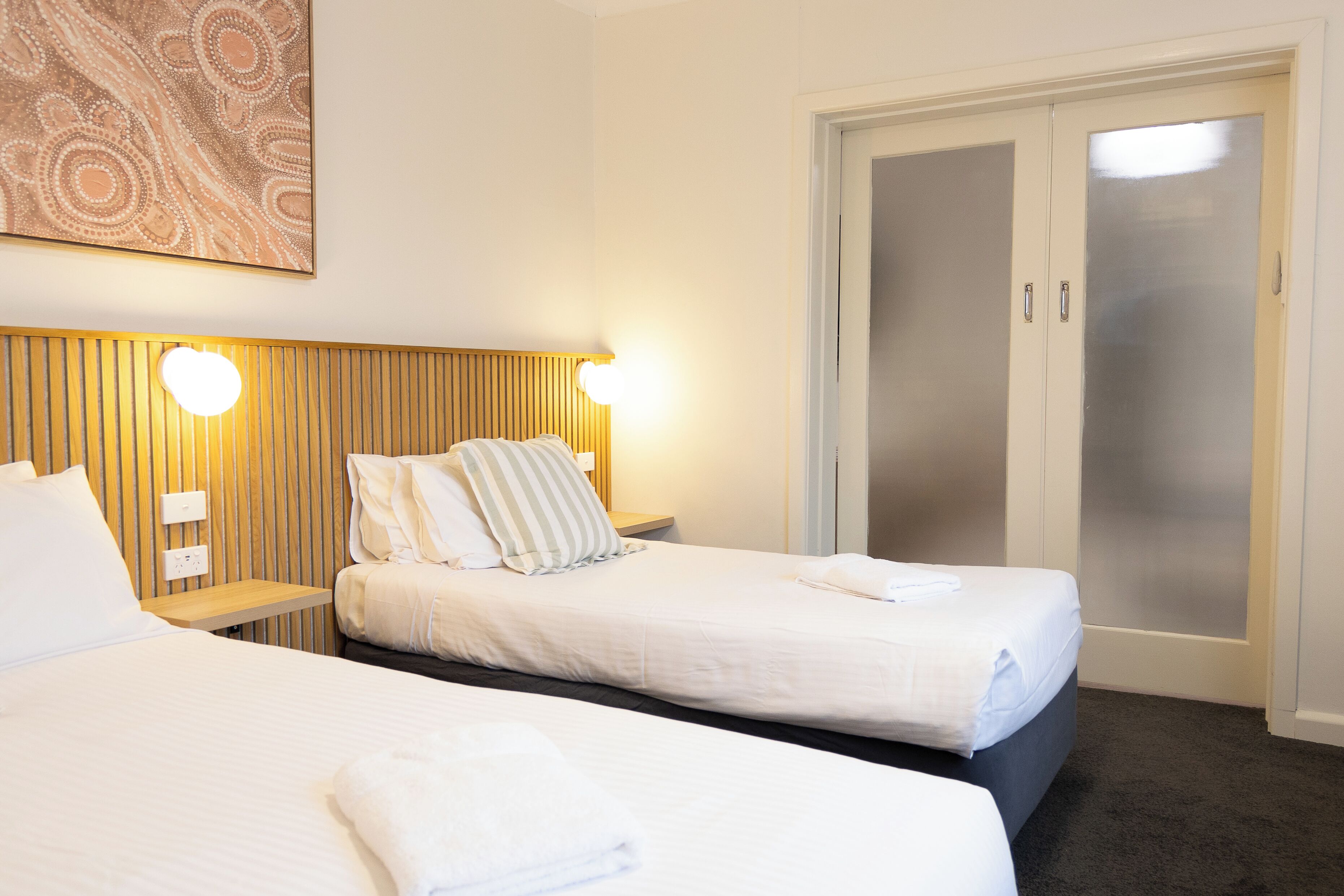 Habitación de hotel con dos camas, cabecero de madera, luz empotrada en la pared y puerta de vidrio satinado.