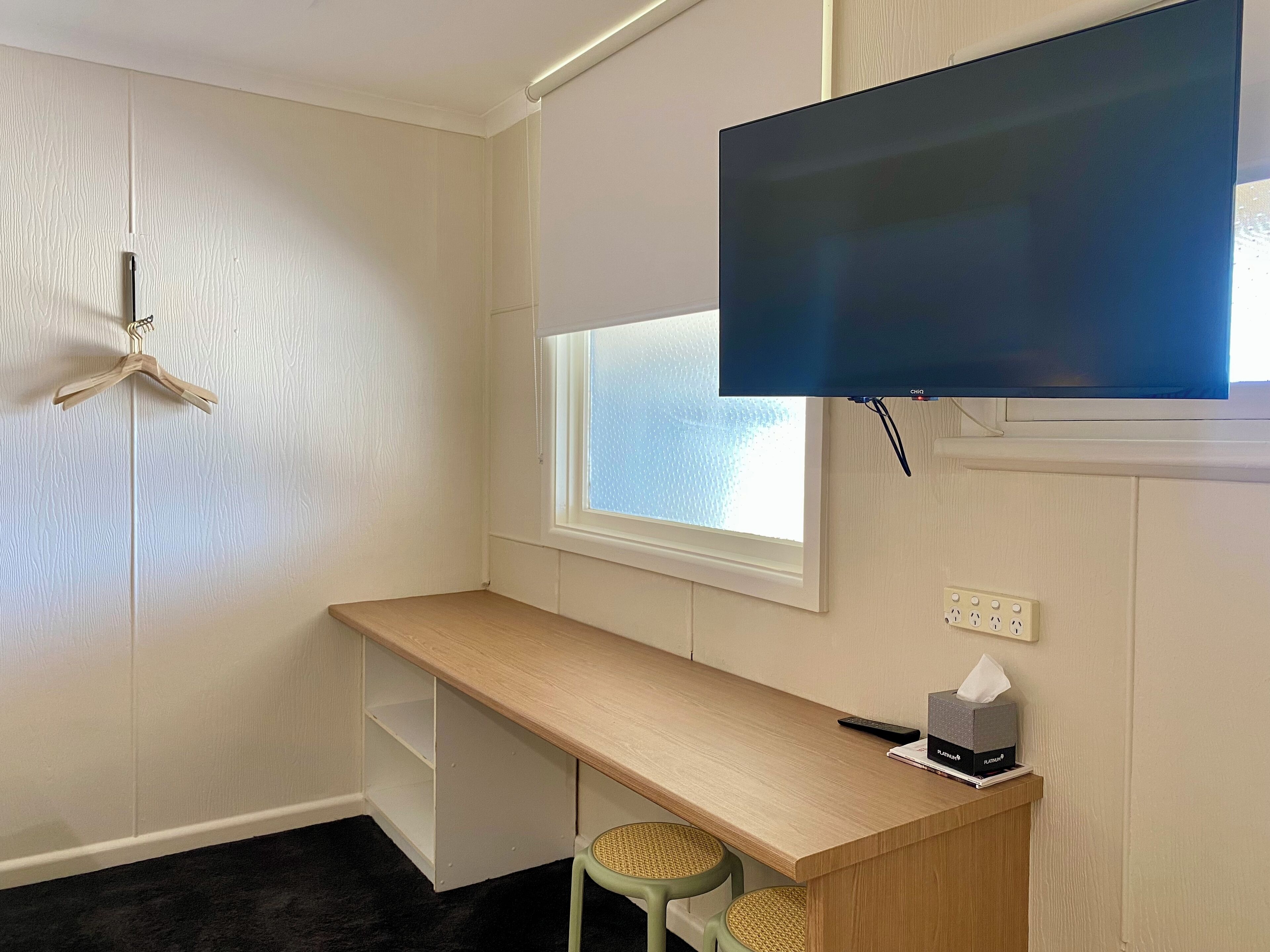 A small, tidy room with a wooden desk, a mounted TV, a window, and a wall-mounted power outlet.