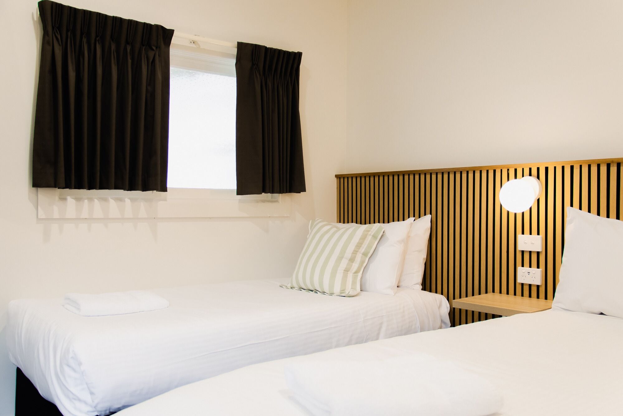 A hotel room with two single beds, a wooden headboard, a bedside table, and a window with black curtains.